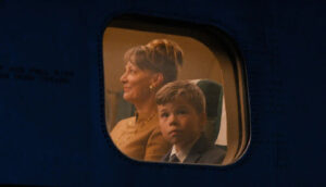 A young boy wearing a suit and tie looks out an airplane window. His mother, wearing a taupe tweed coat, looks straight ahead.