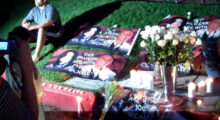 A man wearing a gray t-shirt and black shorts with four white stripes on the side sits on a lawn. Several banners reading "our prayers are with you Michael" are on the ground next to him, as well as white roses and white pillar candles.