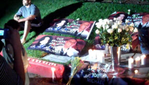 A man wearing a gray t-shirt and black shorts with four white stripes on the side sits on a lawn. Several banners reading "our prayers are with you Michael" are on the ground next to him, as well as white roses and white pillar candles.