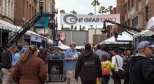 A crowd of people walk down the wide walkway at Universal Studios Lot. A banner hands between buildings that reads "Cine Gear Expo."