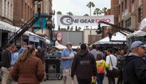A crowd of people walk down the wide walkway at Universal Studios Lot. A banner hands between buildings that reads "Cine Gear Expo."