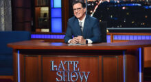 Stephen Colbert, a middle-aged white man with cropped brown hair and black glasses, smiles behind a wood desk that reads "The Late Show." He wears a blue suit, white button-up and gray tie.