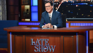 Stephen Colbert, a middle-aged white man with cropped brown hair and black glasses, smiles behind a wood desk that reads "The Late Show." He wears a blue suit, white button-up and gray tie.