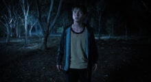 A young man wears a navy hoodie and a beige t-shirt. He stands in the middle of a woodsy area in the dead of night. He looks straight ahead, some blood stains mark his face and shirt.