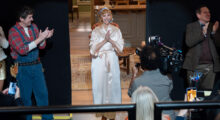 A woman with light copper hair stands on a soundstage and looks out with a big smile at a live studio audience. Her hair is tied up with a blue silk scarf. She wears a white satin robe and holds her hands in a prayer pose. Camera men and boom operators surround her and smile.
