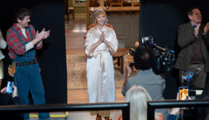 A woman with light copper hair stands on a soundstage and looks out with a big smile at a live studio audience. Her hair is tied up with a blue silk scarf. She wears a white satin robe and holds her hands in a prayer pose. Camera men and boom operators surround her and smile.