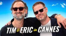 Two middle aged men, both wearing black sunglasses, smile and stand shoulder to shoulder in front of a rich blue sky. Below them text reads" Tim & Eric in Cannes."