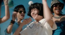 A young Japanese girl wears a white collared shirt with a blue border accent. She smiles and waves her hands in the air. She is surrounded by other people who are also jumping and waving. A blue sky is visible behind them.