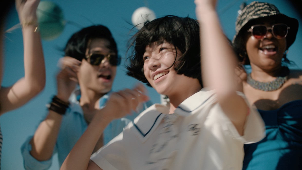 A young Japanese girl wears a white collared shirt with a blue border accent. She smiles and waves her hands in the air. She is surrounded by other people who are also jumping and waving. A blue sky is visible behind them.